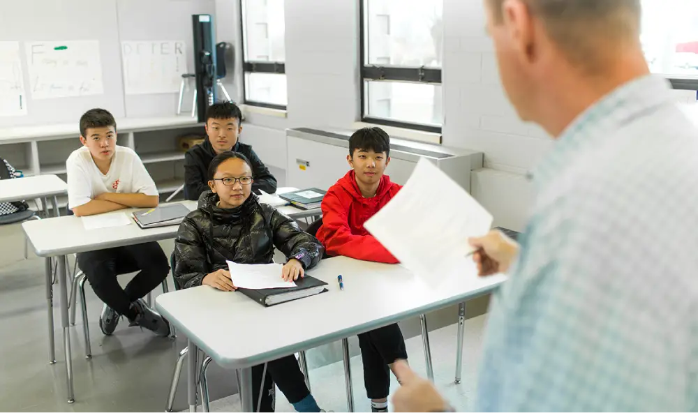 A teacher is speaking to four students seated at desks in a classroom.