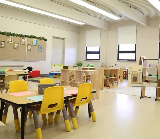 An empty classroom with small tables, yellow chairs, and shelves with toys and learning materials.