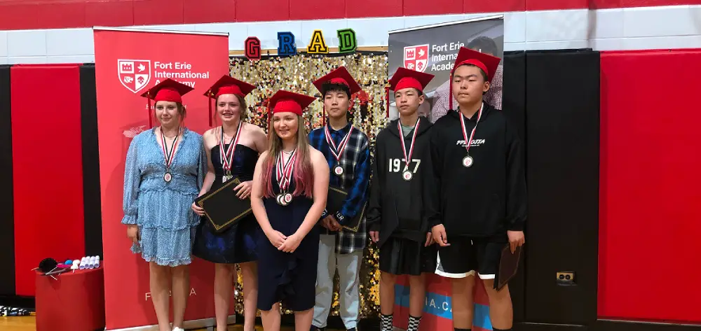 Six students in red graduation caps and medals stand proudly on a stage with a "GRAD" sign.