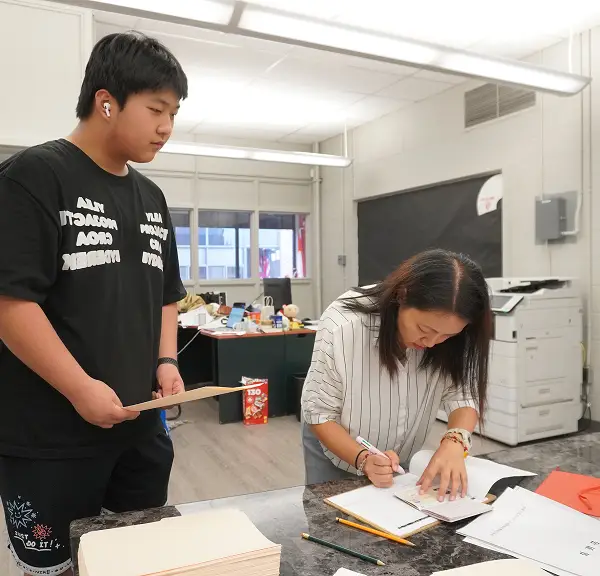 A student hands papers to a teacher who is writing at a desk in a classroom.