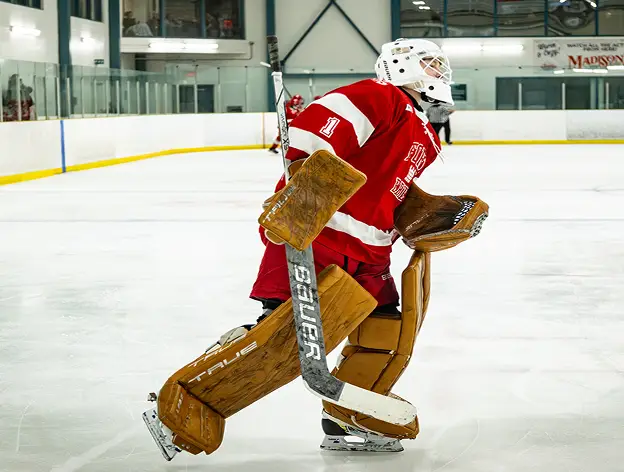 A hockey goalie in red gear stands on the ice, holding a stick with focus.