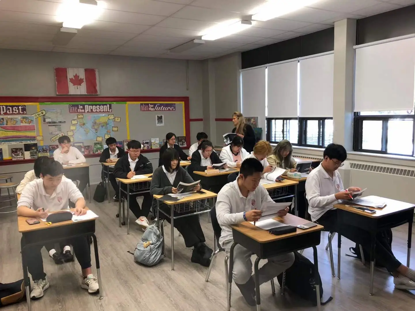 Students in a classroom sitting at desks and reading textbooks with a teacher standing nearby.