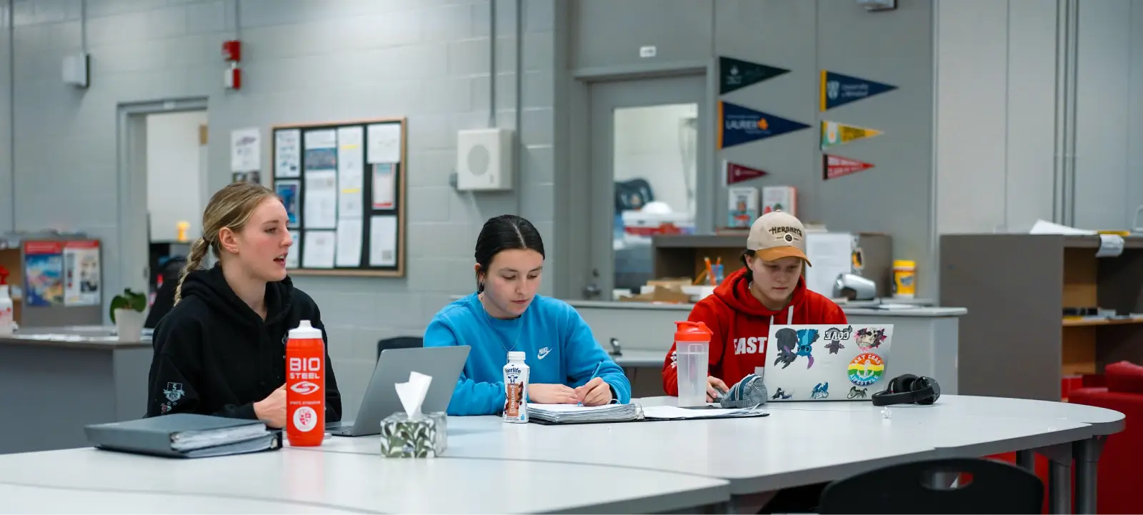 Three people sitting at a table, working on laptops and taking notes in a classroom setting.