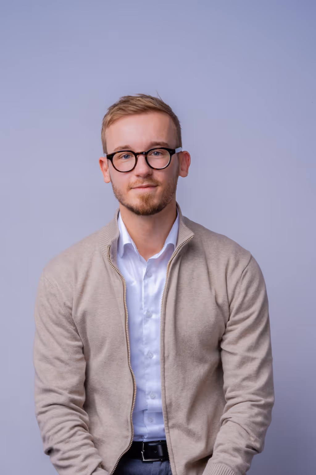 Young man with short light brown hair, wearing round black glasses, a beige zip-up sweater, and a white collared shirt, sitting against a plain light gray background.