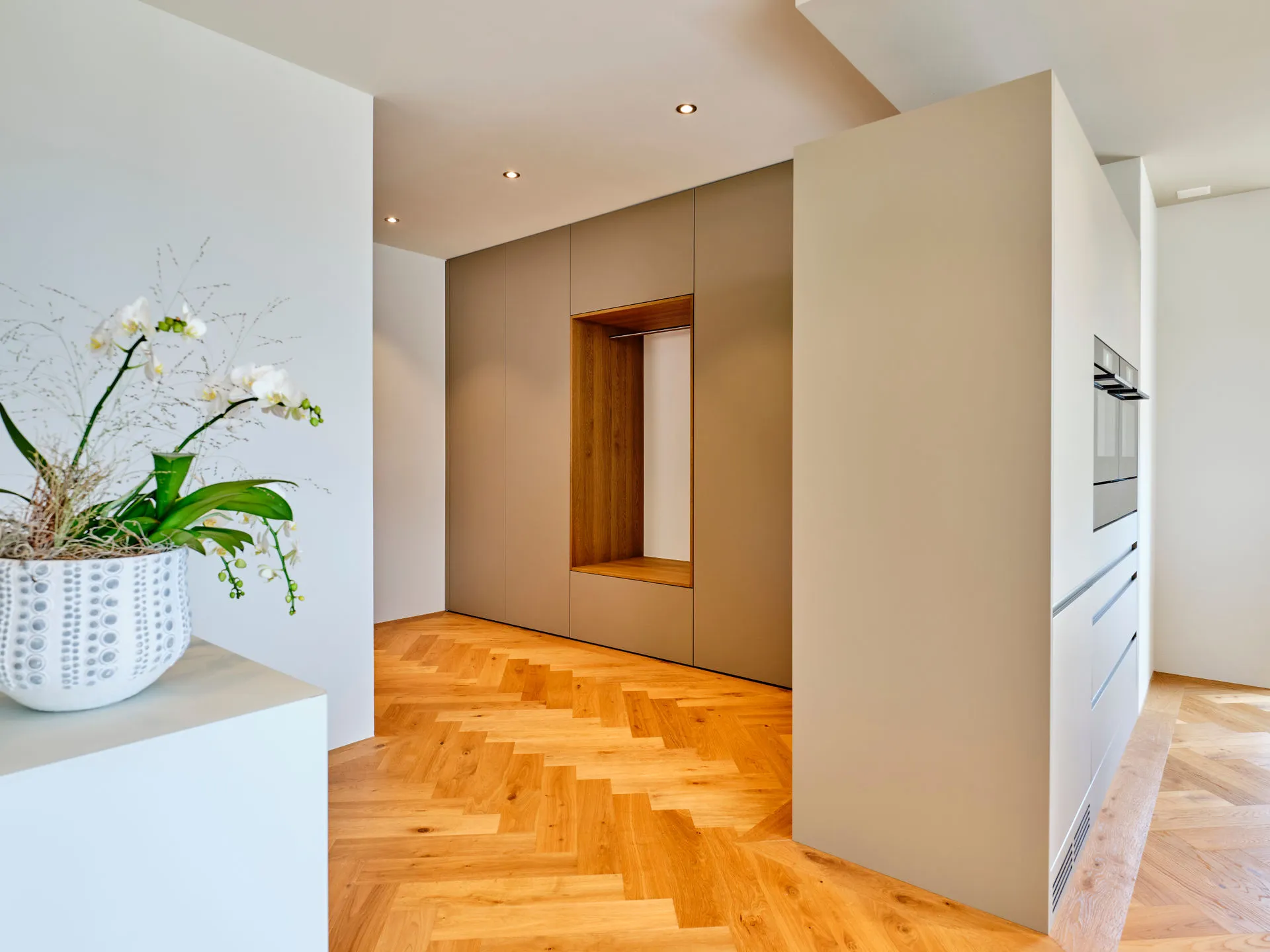 Heller Eingangsbereich mit hölzernen Fischgrätparkett, eingebauter Garderobe in grauen Einbauschränken und weissen Wänden.