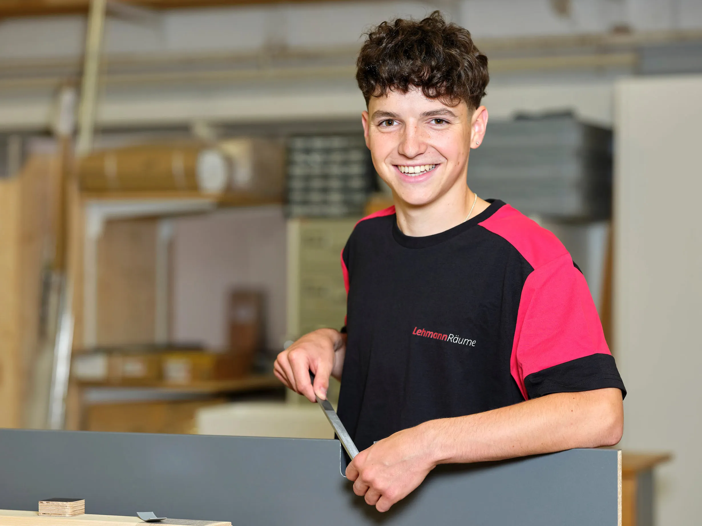 Junger Mann in schwarzem und rotem Lehmann Räume T-Shirt arbeitet in einer Werkstatt und feilt an einem grauen Werkstück.