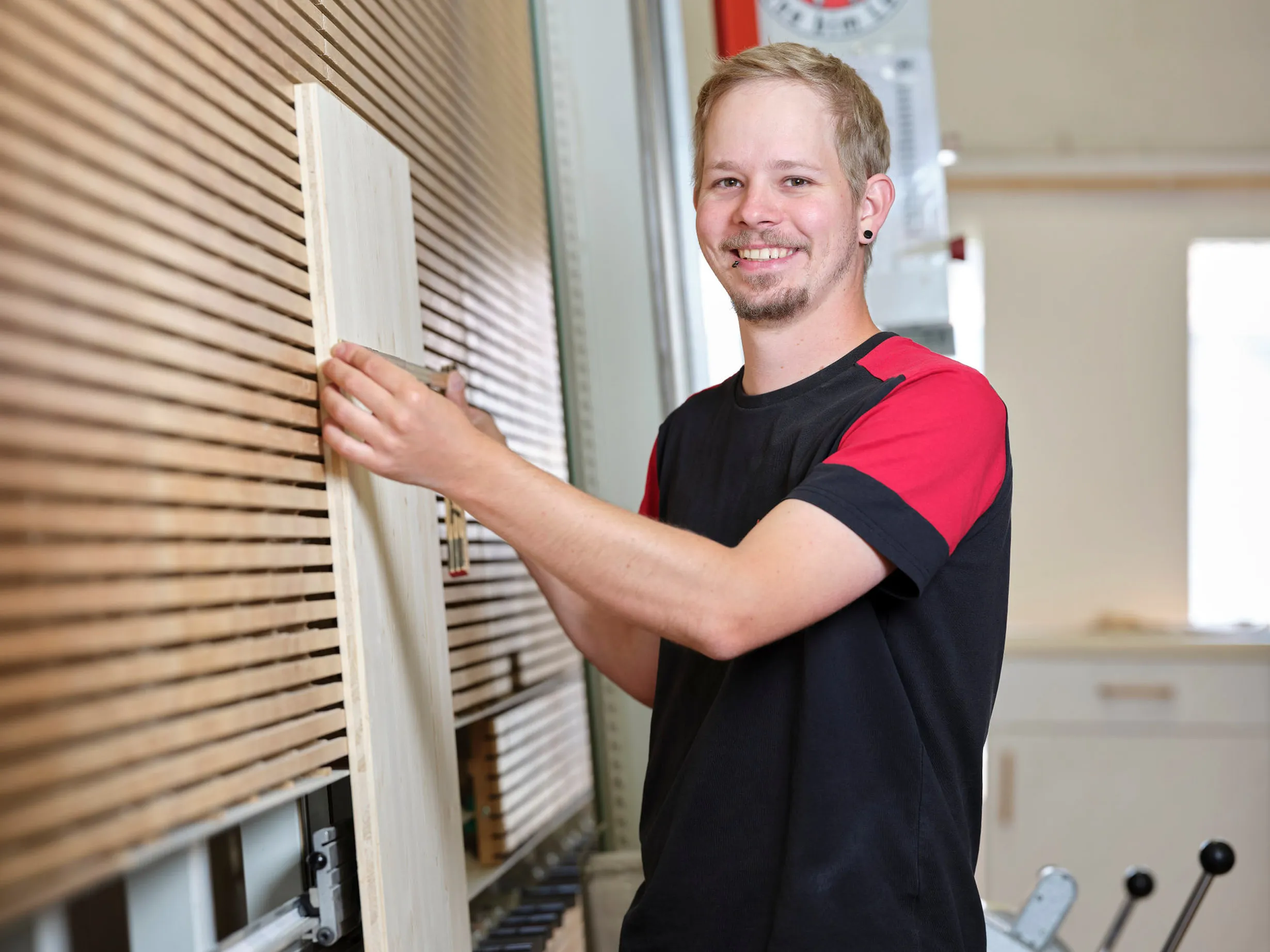 Junger Mann mit blondem Haar und rotschwarzem T-Shirt hält ein Holzbrett vor einem gestapelten Holzregal in einer Werkstatt.