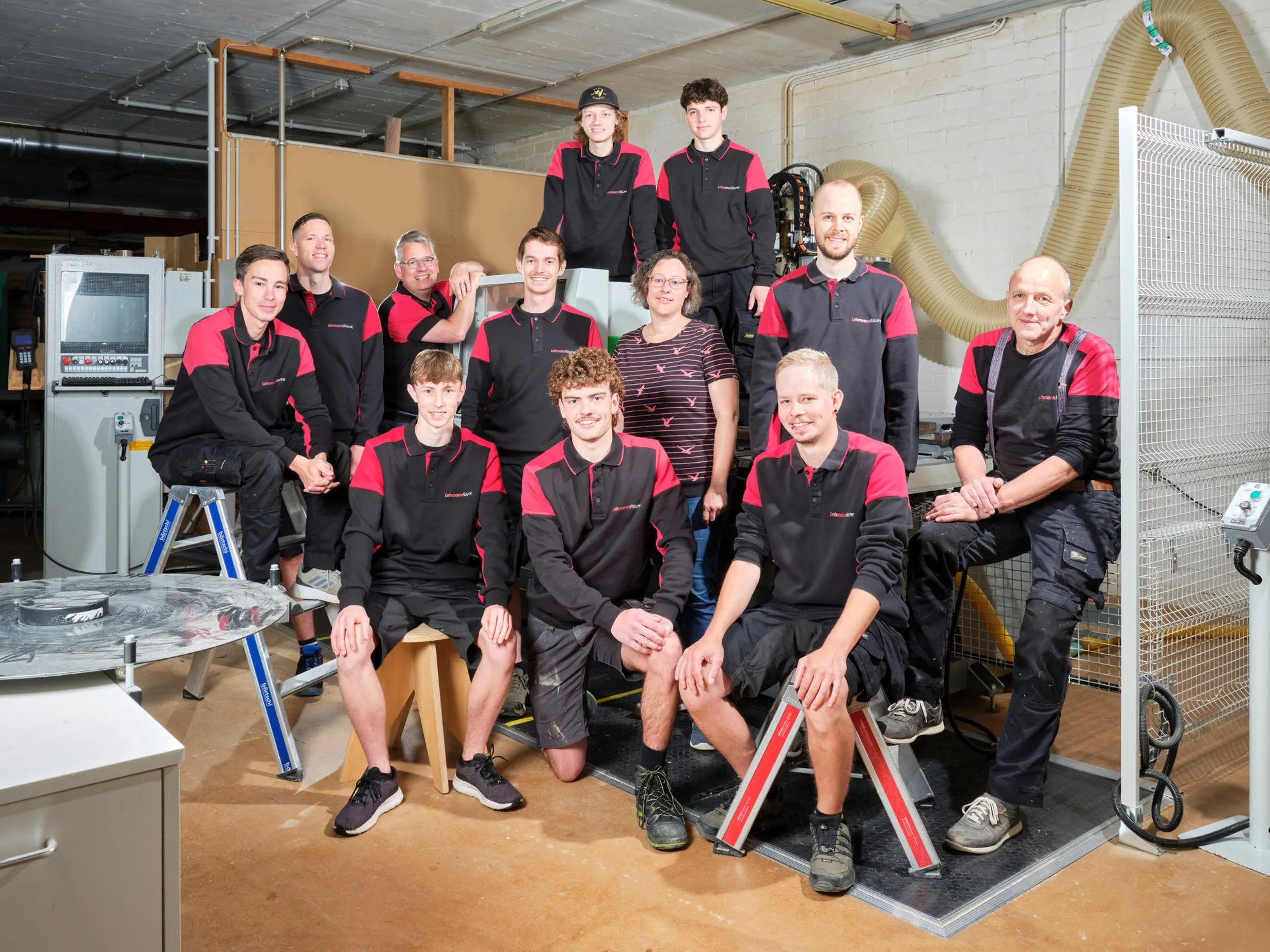 Das Team von zwölf Personen in einer Werkstatt, die meisten tragen schwarze und rote Arbeitskleidung mit dem Schriftzug 'LehmannRäume'.