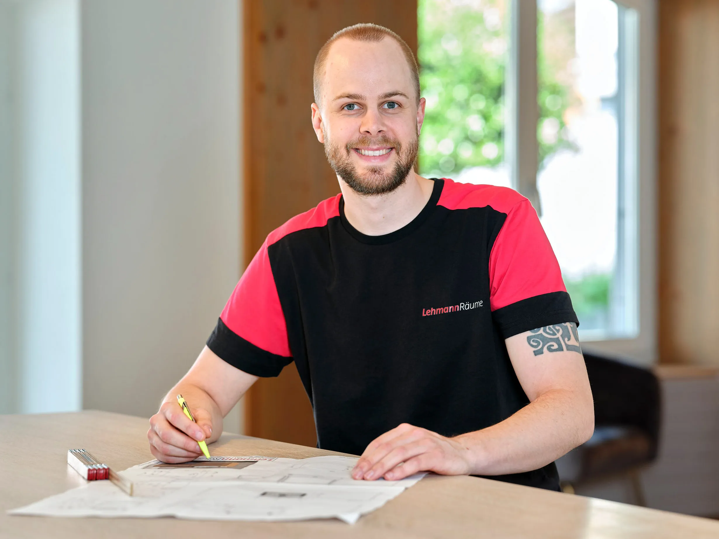 Lächelnder Mann mit Bart und Tätowierung zeichnet auf einem Tisch an einer Bauzeichnung.