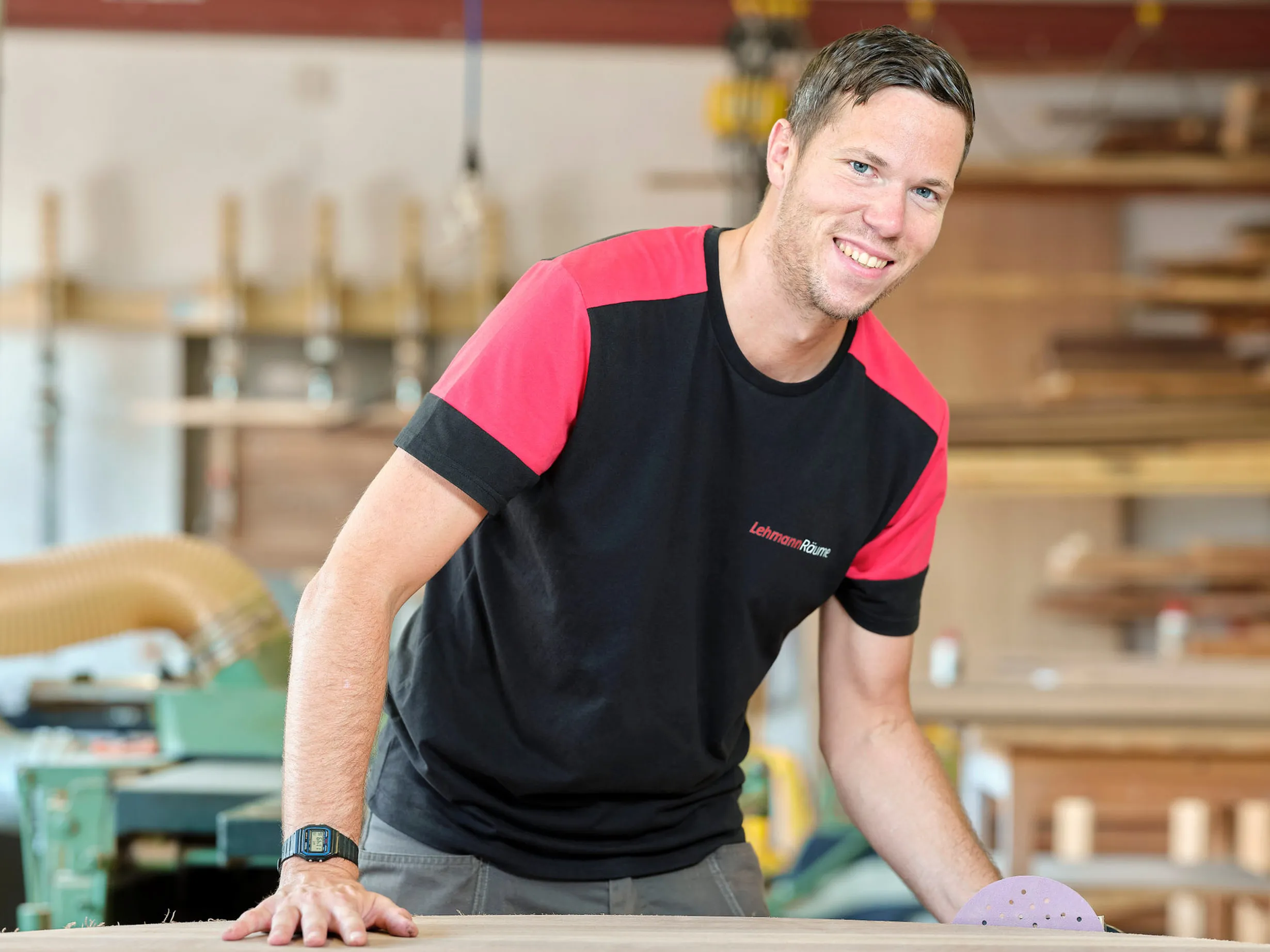 Lächelnder Mann in Werkstatt lehnt sich auf eine Holzplatte, trägt T-Shirt mit Lehmann Räume Logo.