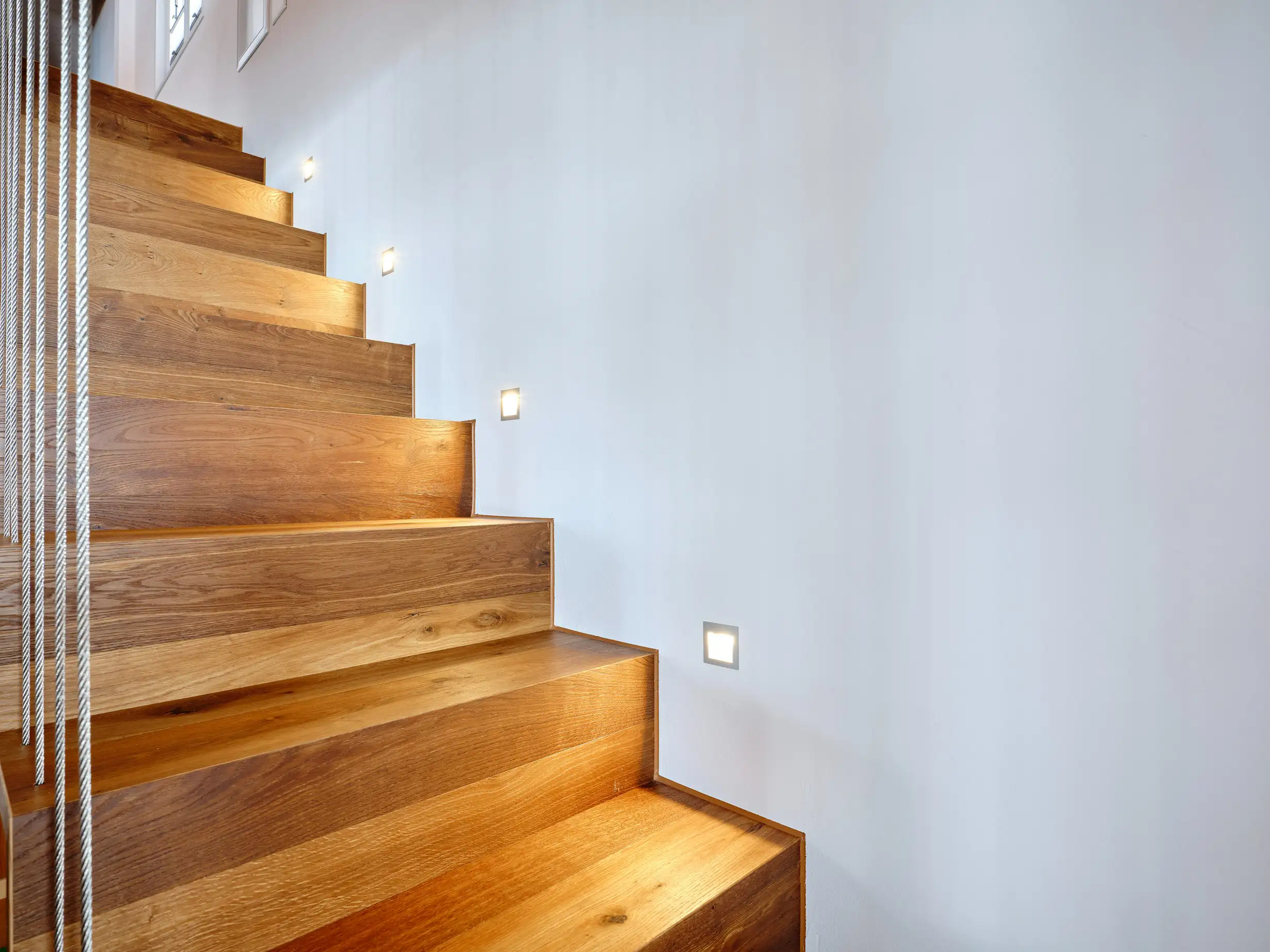 Moderner Holztreppenaufgang mit eingebauten Wandlichtern an weissem Treppenhaus.