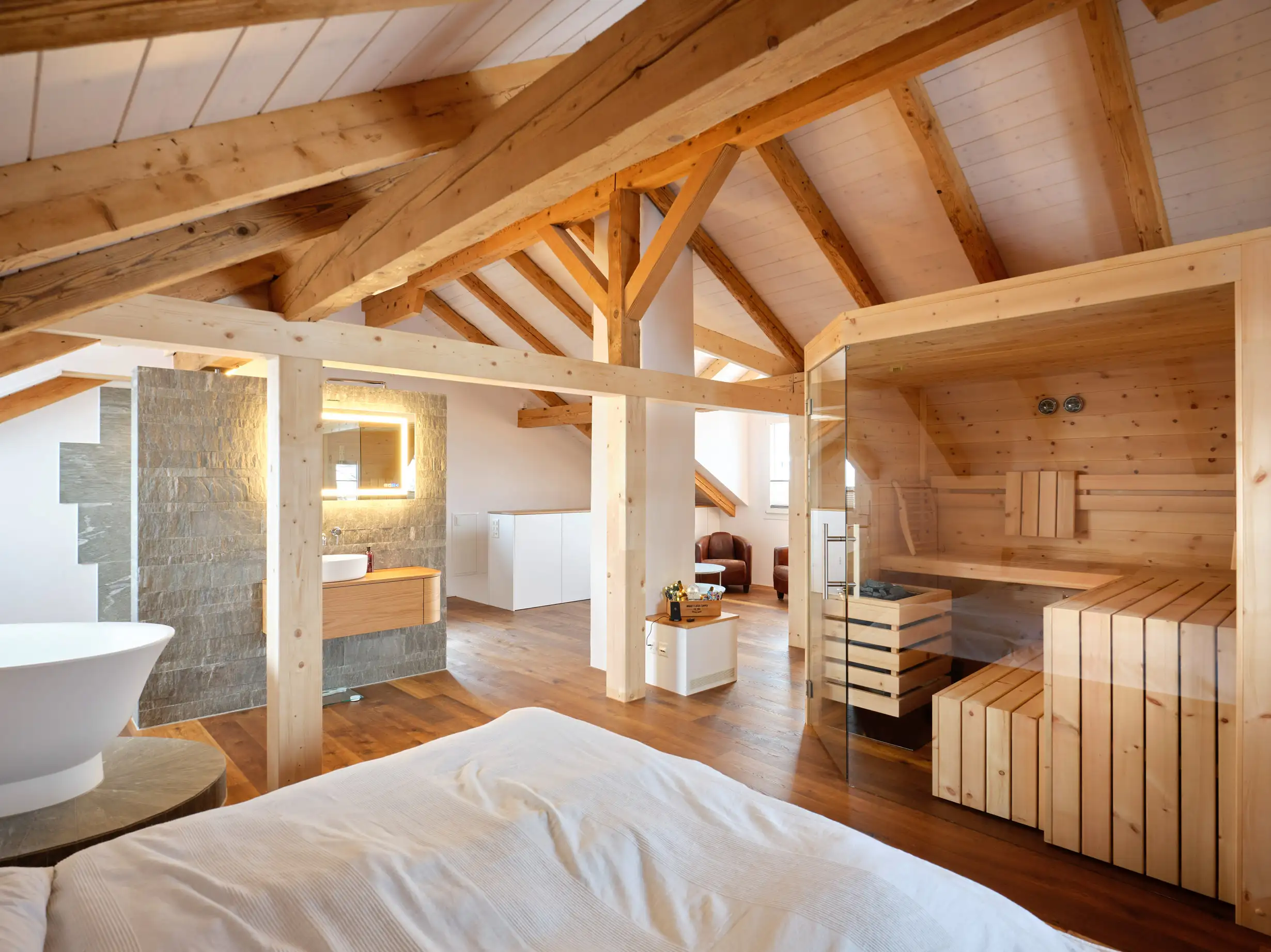 Modernes Dachzimmer mit sichtbaren Holzträgern, freistehender Badewanne, Waschtisch mit beleuchtetem Spiegel und Sauna mit Glastür.
