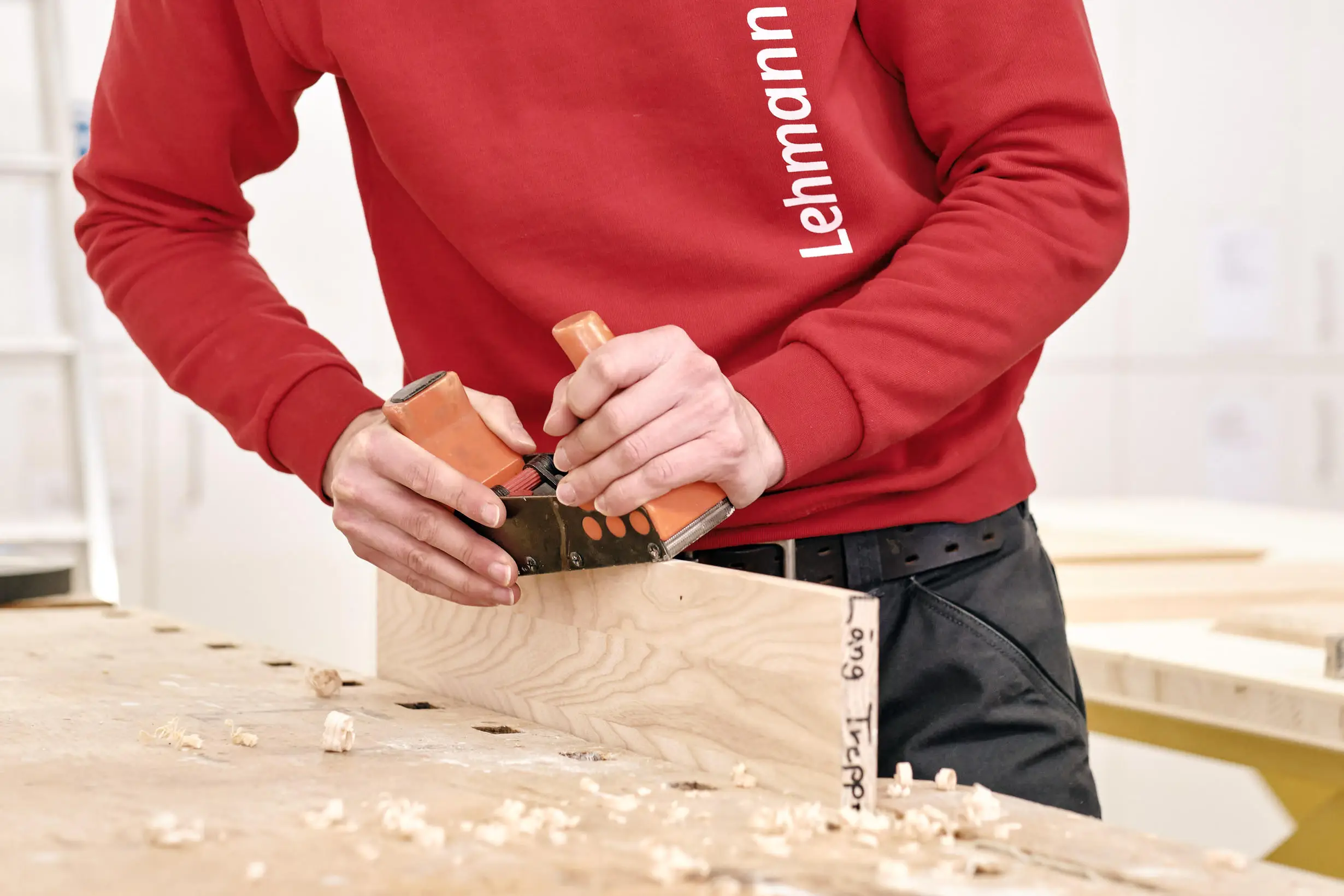 Eine Person in rotem Pullover benutzt ein Holzhobelgerät an einem Holzbrett mit Holzspänen auf der Werkbank.