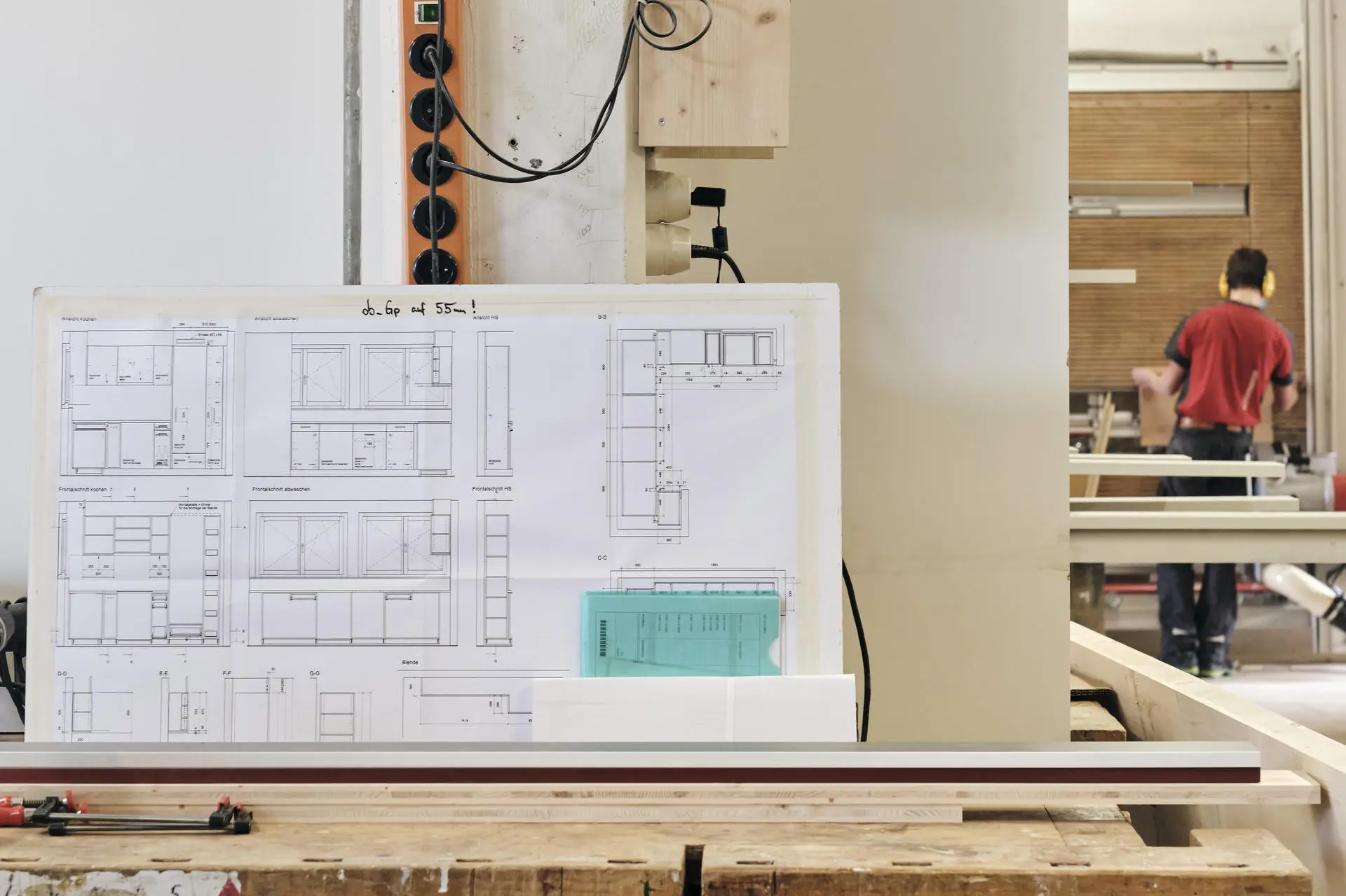 Baupläne auf einer Werkbank in einer Schreinerei, im Hintergrund arbeitet eine Person mit Gehörschutz.
