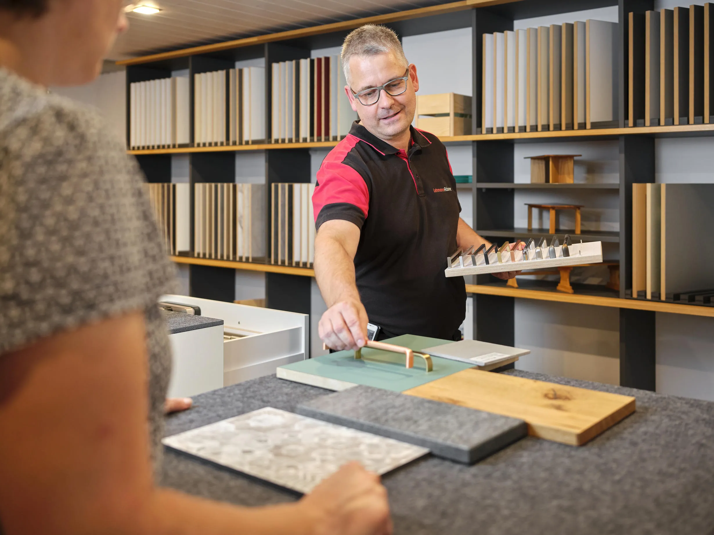 Ein Mann zeigt einer Kundin verschiedene Möbelgriffe auf Musterplatten in einem Schauraum mit Regalen voller Holz- und Oberflächenproben.