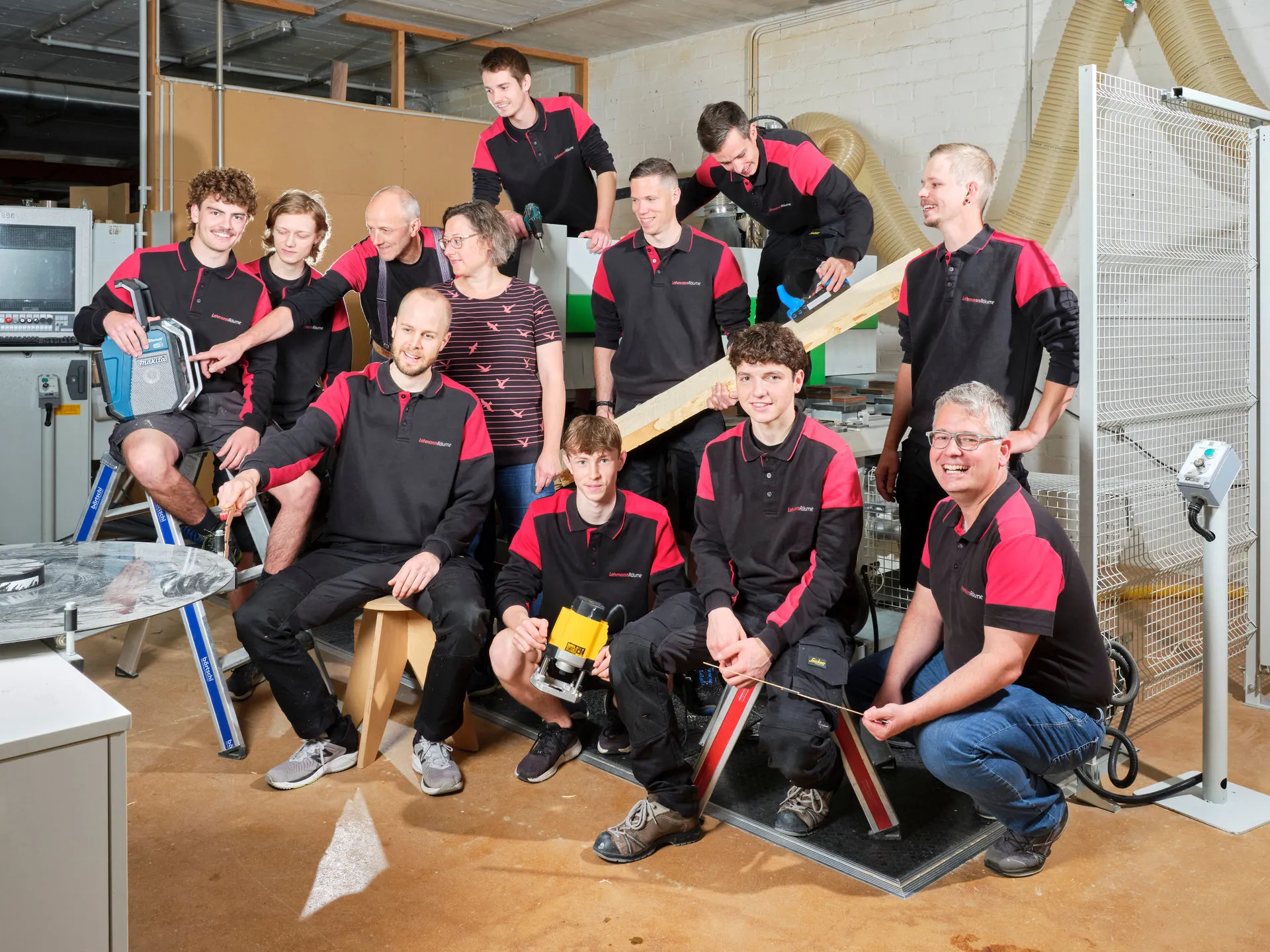 Gruppenbild von zwölf Personen in Werkstatt, die rote und schwarze Firmenpolos tragen und Holzwerkzeuge und Maschinen benutzen.