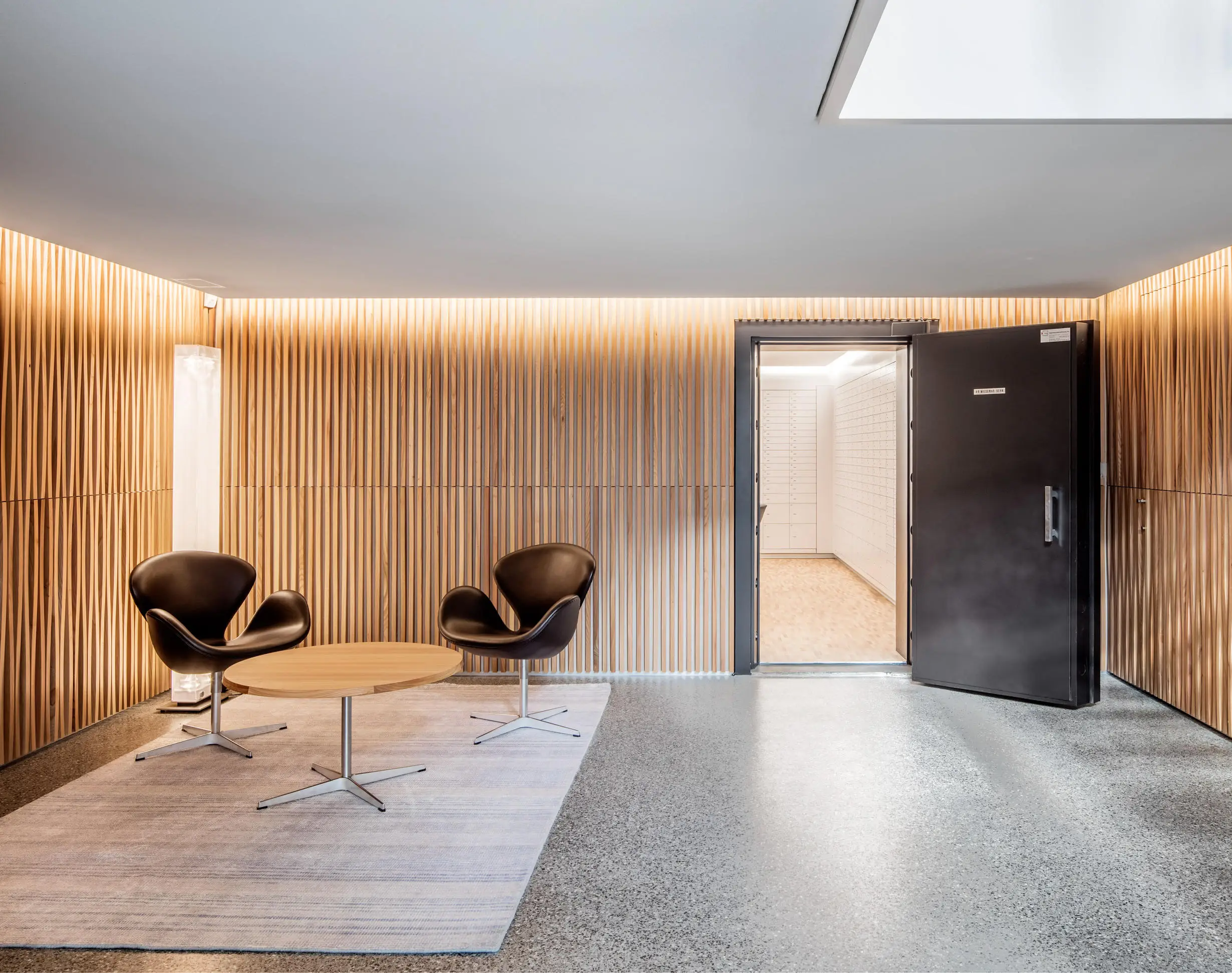 Modernes Wartezimmer mit zwei schwarzen Sesseln, einem runden Holztisch, einer strukturierten Holzverkleidung und einer offenen schwarzen Tür zum hellen Raum mit vielen Schliessfächern.