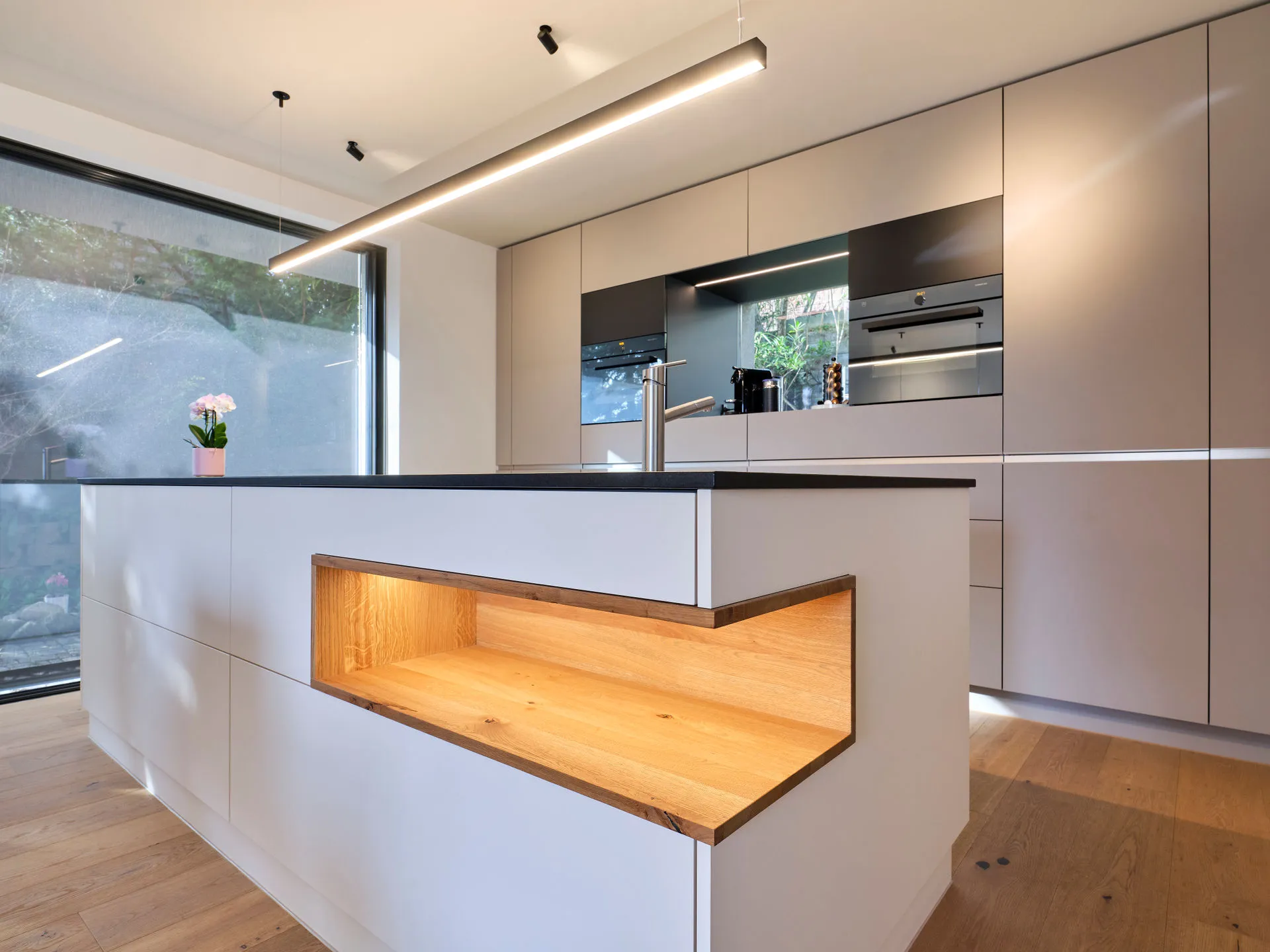 Moderne Küche mit Insel, eingebautem Backofen und Fenster mit Blick ins Grüne.