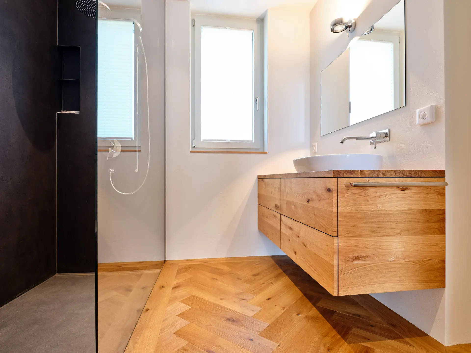 Modernes Badezimmer mit hölzernem Waschbeckenunterschrank, rundem Aufsatzwaschbecken, großem Spiegel und begehbarer Dusche mit schwarzer Wand.