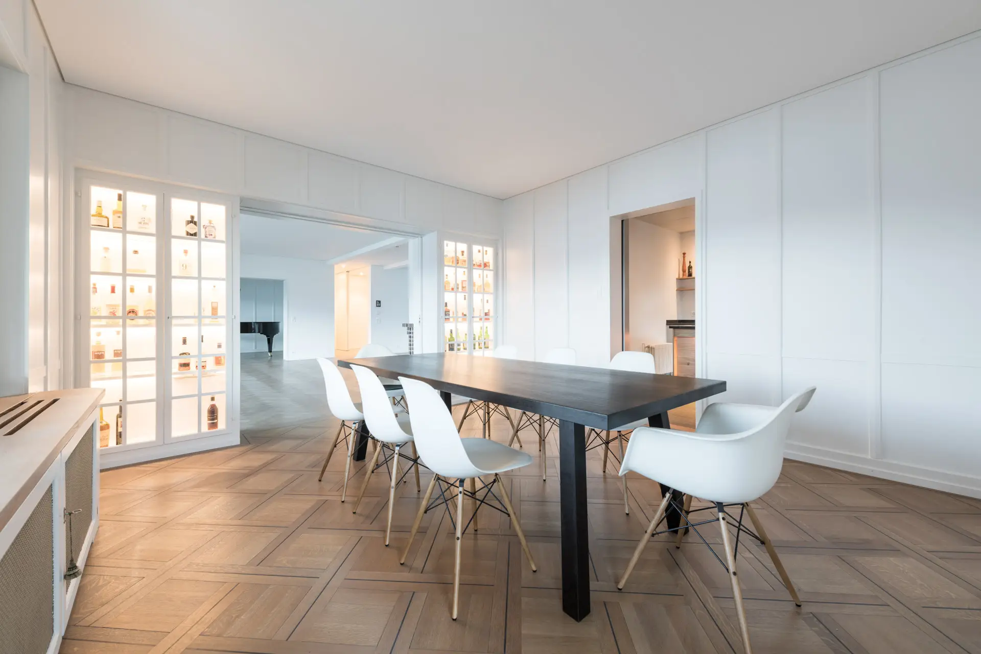 Moderner Essbereich mit dunklem rechteckigem Tisch und acht weißen Stühlen auf Holzparkettboden, umgeben von weißen Wänden mit beleuchteten Vitrinen.
