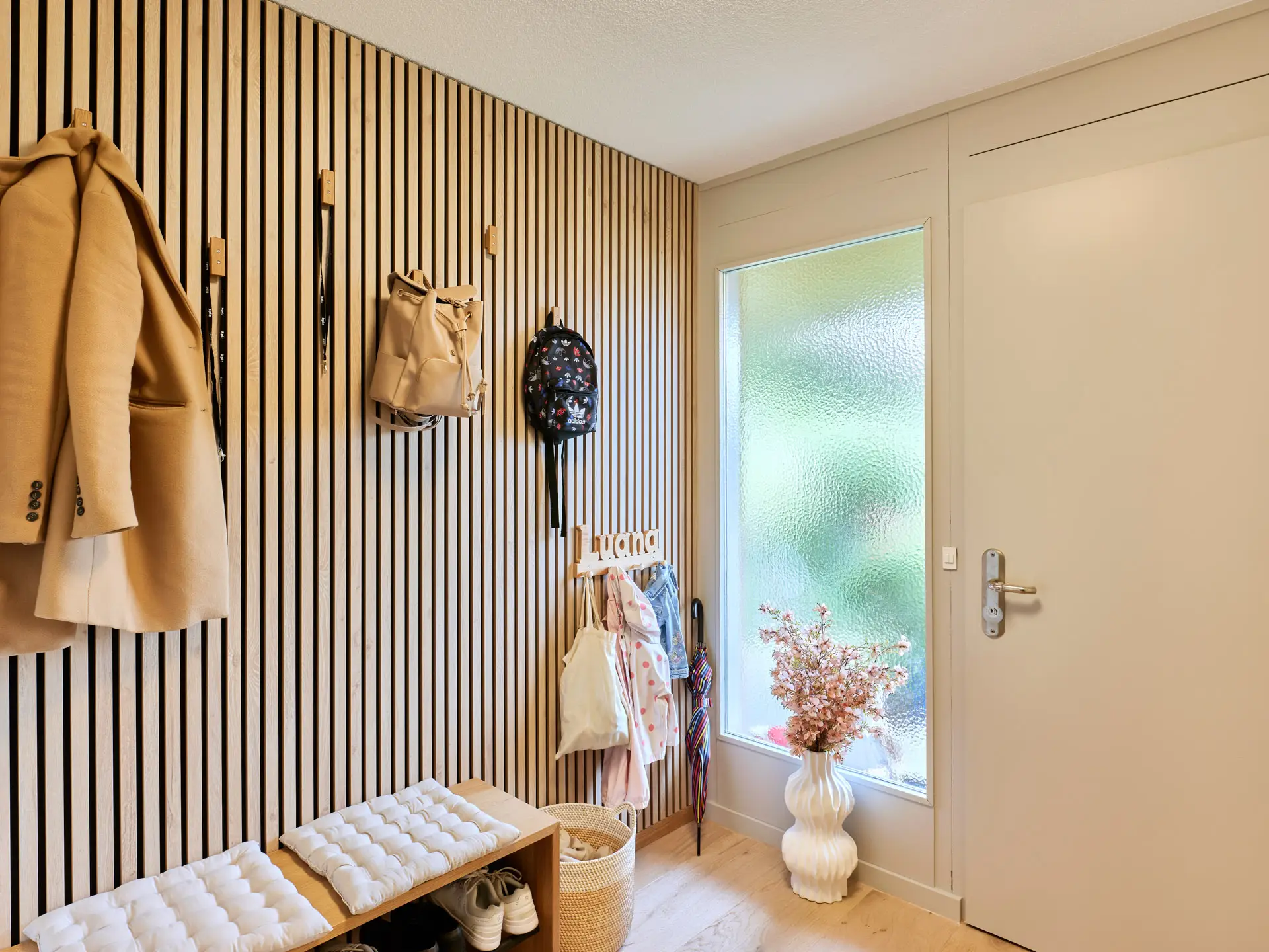 Heller Eingangsbereich mit Holzvertäfelung, Sitzbank mit Kissen, hängenden Jacken, Rucksäcken und einem Blumenstrauss in Vase neben einer Tür mit satiniertem Fenster.