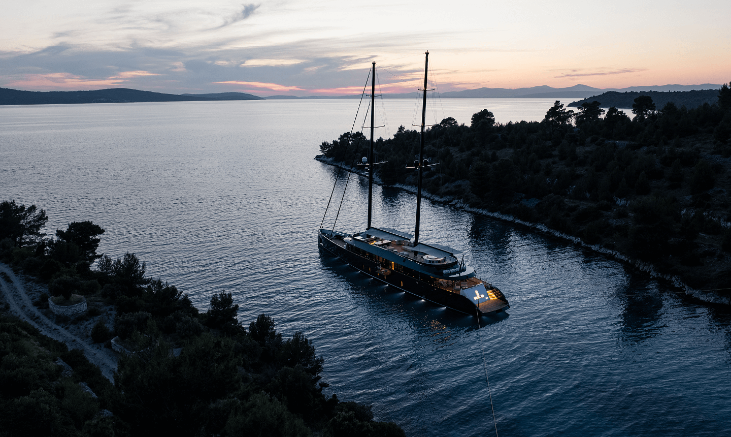 A luxury yacht anchored in calm waters at sunset, surrounded by trees and a mountainous coastline.
