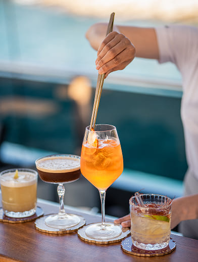 Hand using long tongs to stir an orange cocktail with ice on a table alongside three other mixed drinks.