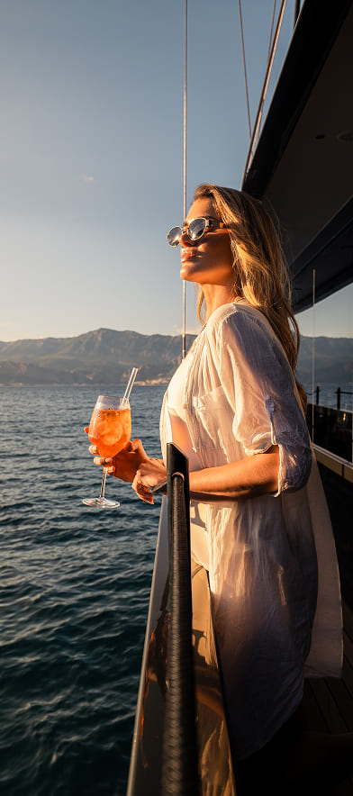 Woman in sunglasses and a sheer white cover-up holding a cocktail, standing on a yacht overlooking the ocean at sunset.