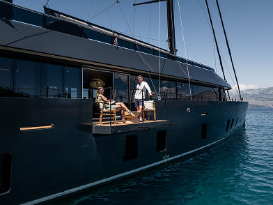 Two people relaxing on chairs on the side deck of a large black yacht on calm blue water.