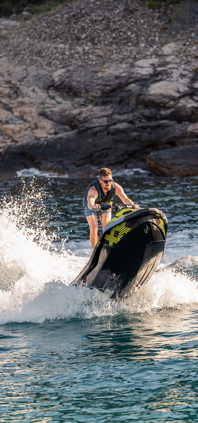 Man wearing sunglasses riding a jet ski over water with rocky shore in the background.