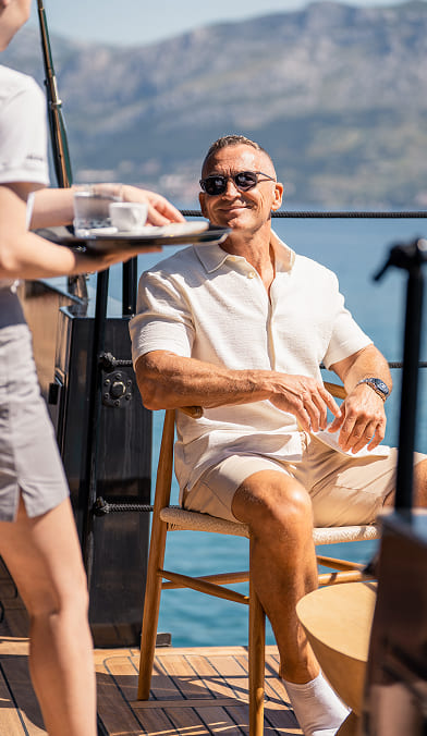 Man wearing sunglasses and light summer clothes sitting on a wooden chair on a boat, receiving a tray with a cup and glass from a server.