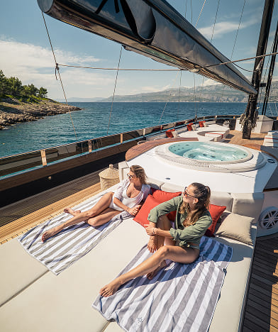 Two women relaxing on striped towels on a yacht deck with ocean and coastline in the background.
