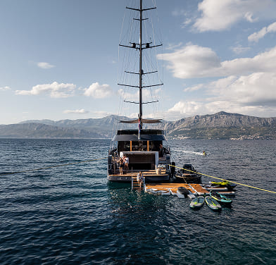 Sailboat anchored on calm water with paddleboards and mountains in the background under a partly cloudy sky.