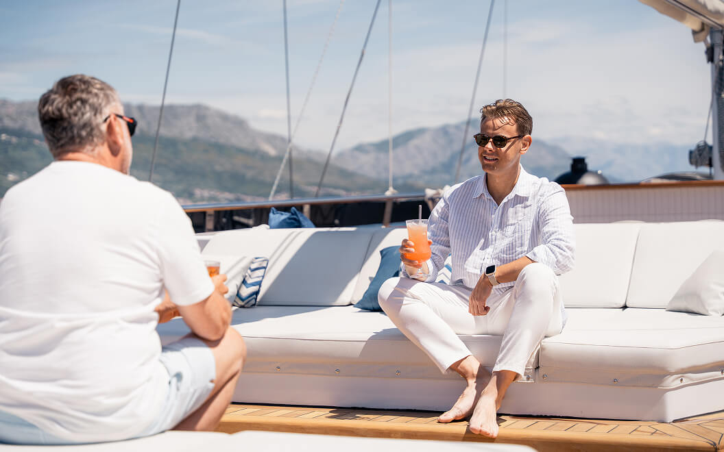 Two men relaxing and drinking cocktails on a yacht with mountains in the background.