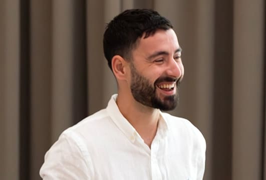 Smiling man with dark hair and beard wearing a white shirt against a blurred brown background.