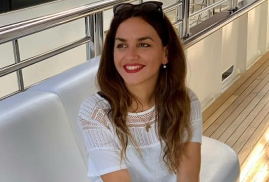 Smiling woman with long brown hair sitting on a white bench wearing a white top and sunglasses on her head.