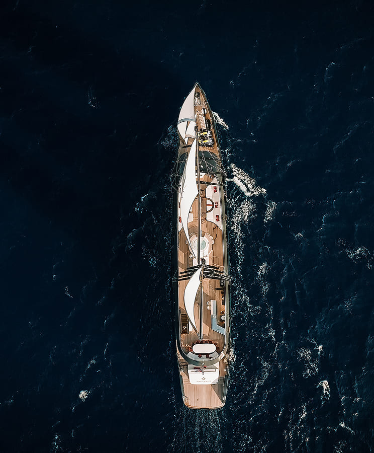 Aerial view of a sailing yacht with white sails navigating through dark blue ocean water.