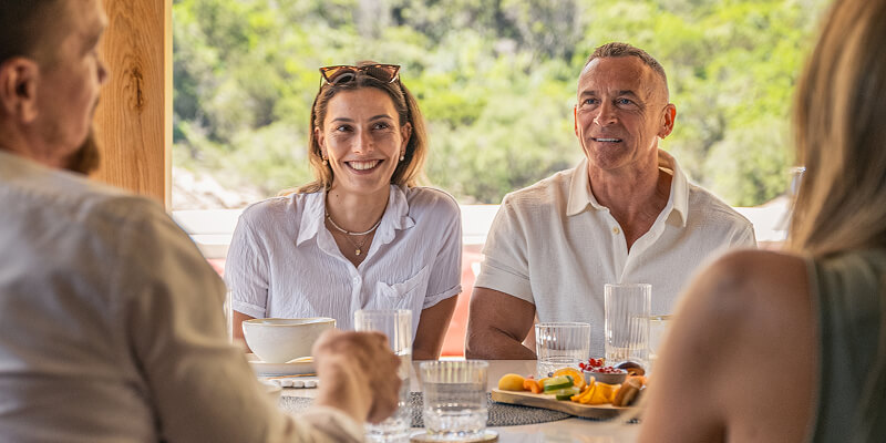 Two people smiling and sitting at a table set with glasses and food, engaged in conversation with two others.