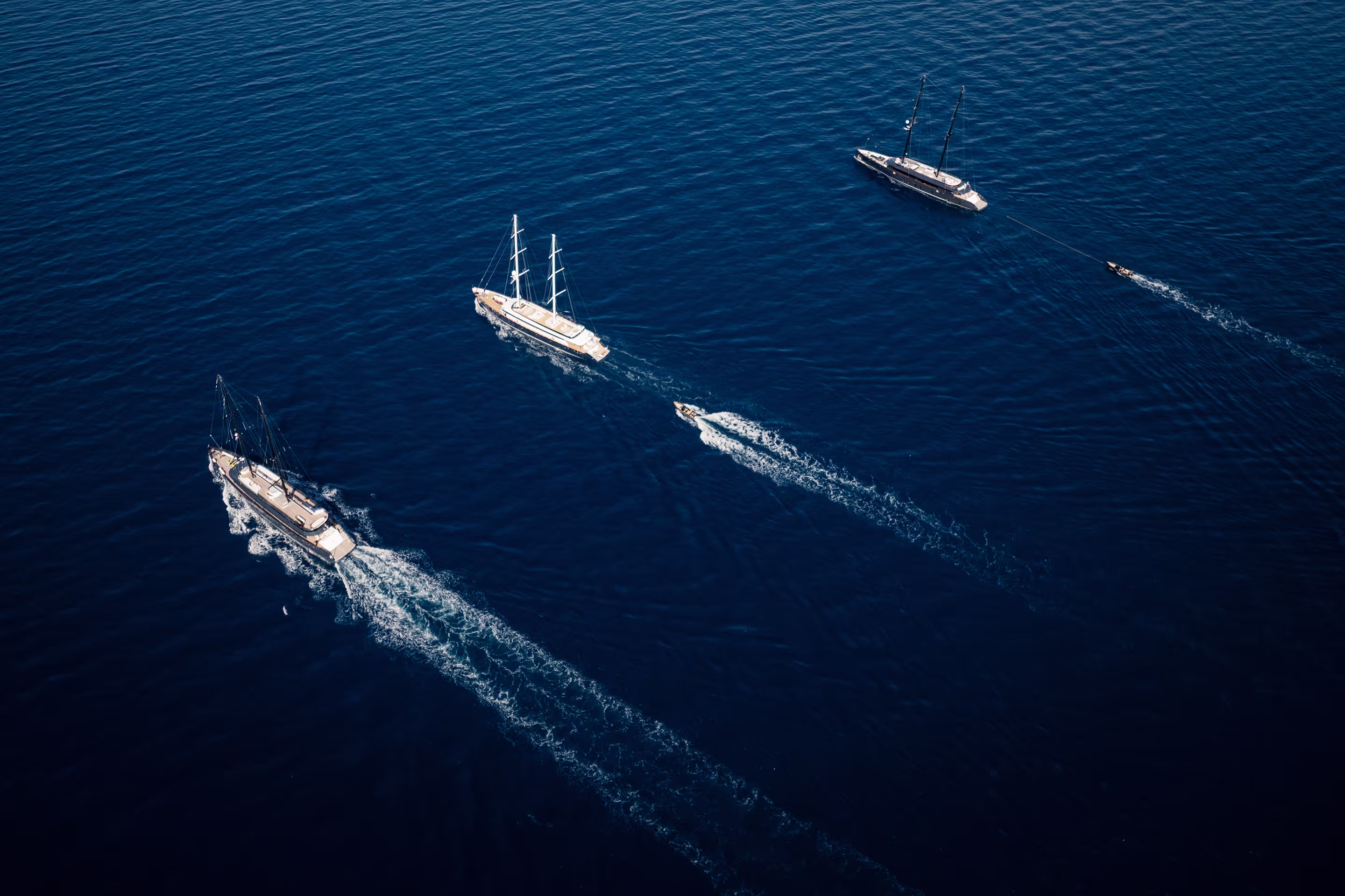Aerial view of three sailboats on dark blue ocean water, each accompanied by a small motorboat creating white wakes.