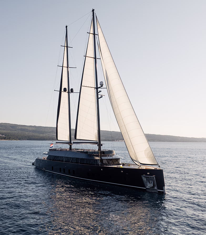 Large modern sailing yacht with black hull and three white sails cruising on calm water near a coastline.