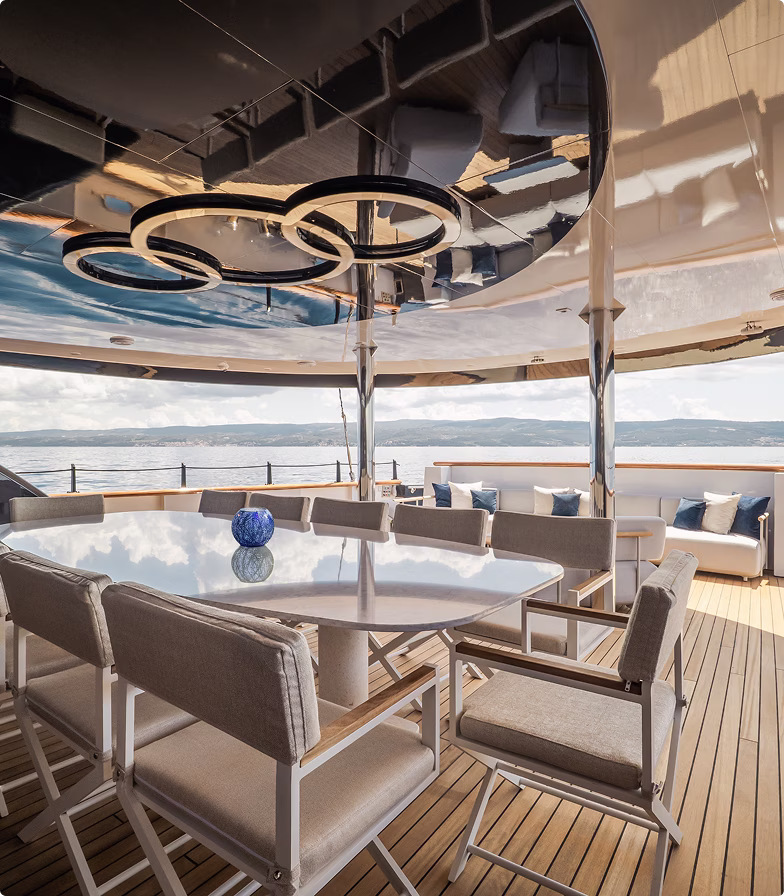 Outdoor dining area on a yacht with beige cushioned chairs around a glossy table and a view of the sea and distant hills.