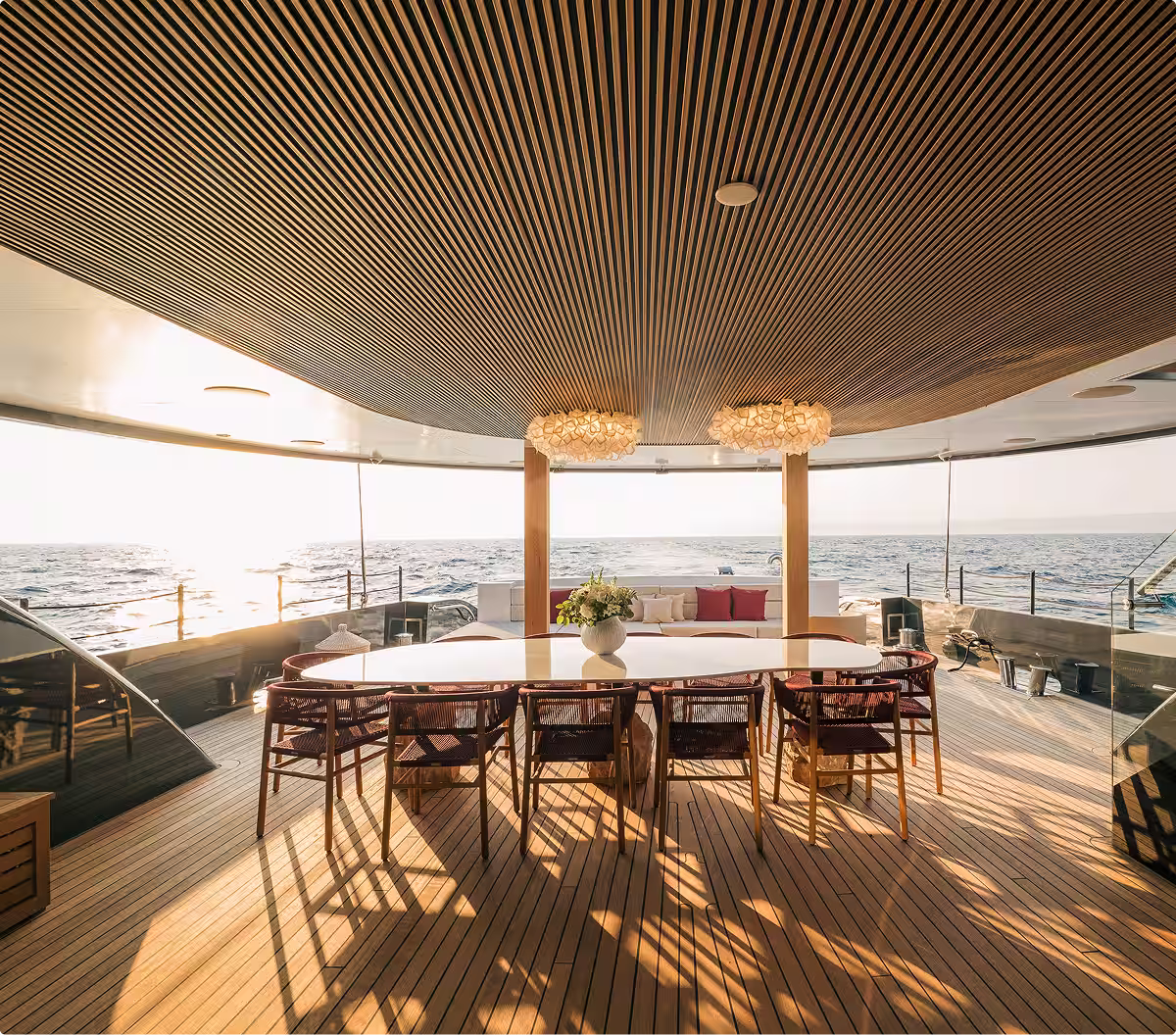 Outdoor dining area on a yacht with wooden flooring and ceiling, a large table with chairs, and ocean view at sunset.