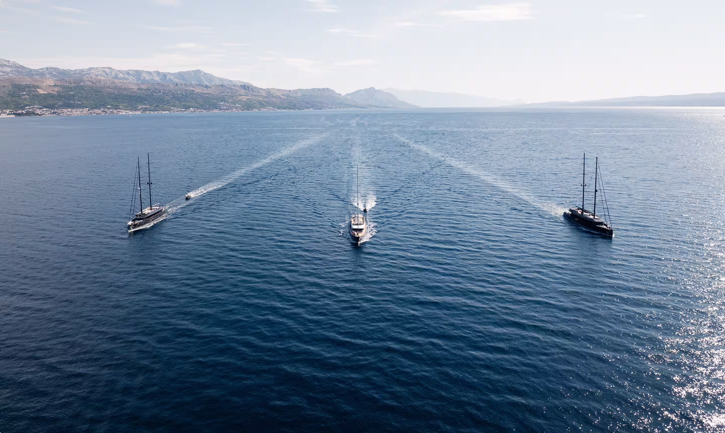 Three sailboats sailing on calm sea water with mountainous coastline in the background.