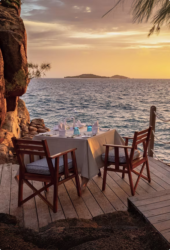 Table set for two with napkins and glasses on a wooden deck overlooking the ocean at sunset.