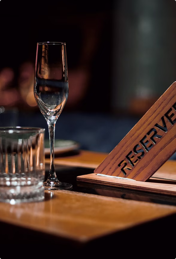 Empty reserved table with a champagne flute, a tumbler glass, and a wooden 'Reserved' sign on a wooden surface in low lighting.