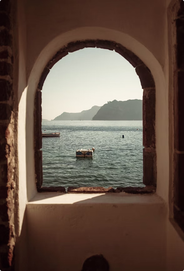 View of calm sea with a small boat and distant mountains framed by an arched stone window.