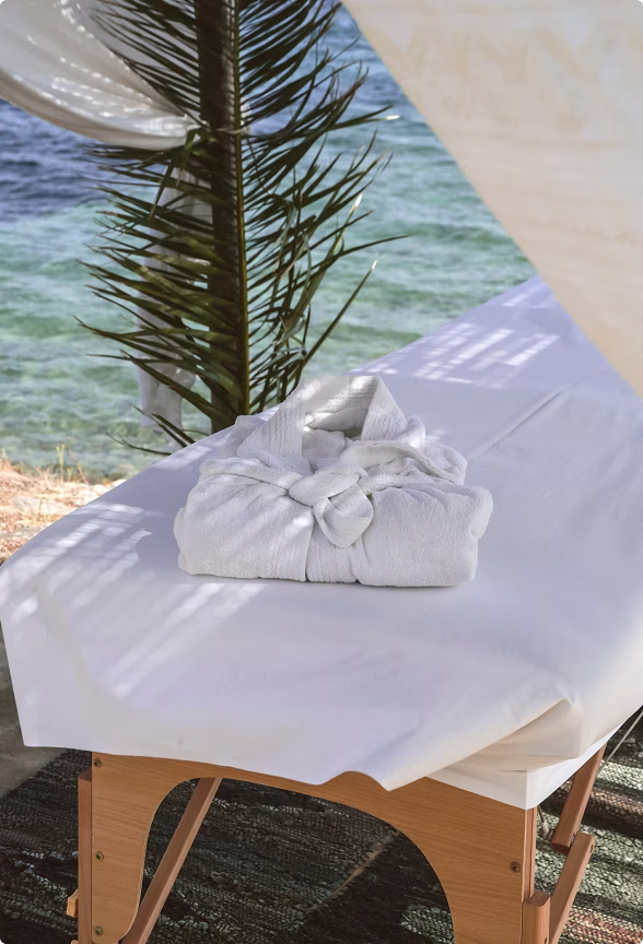 White bathrobe neatly folded on a massage table with a view of the sea and palm leaf in the background.