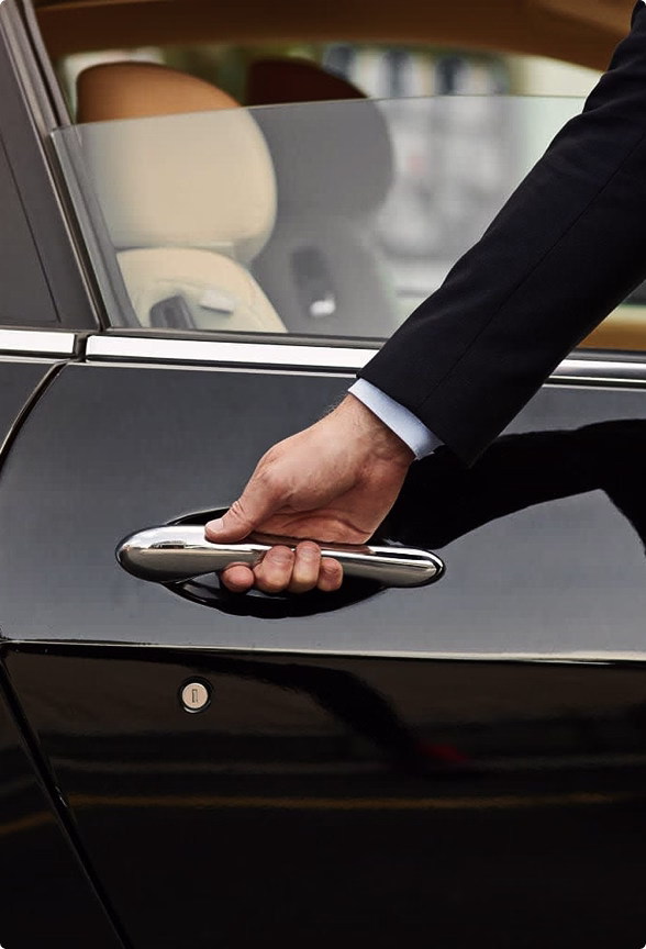 A person in a suit opening the door handle of a black luxury car with beige leather interior.