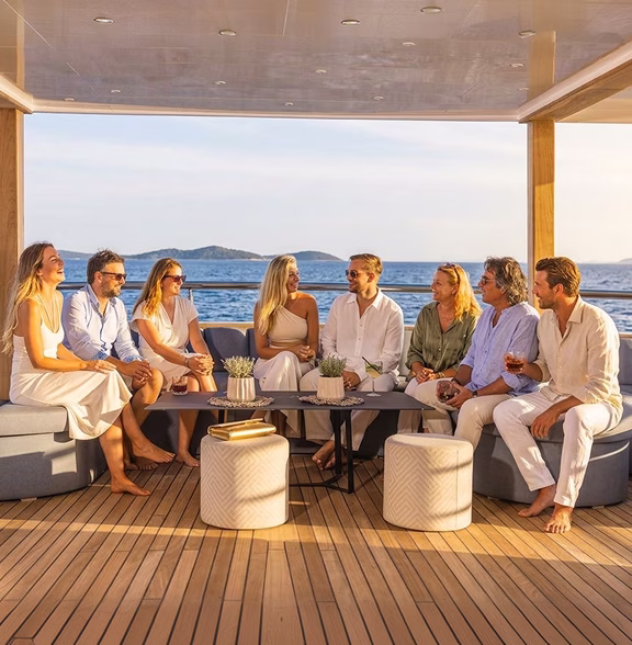 Group of seven people sitting on a yacht deck lounge, enjoying drinks and smiling with the sea and islands in the background.