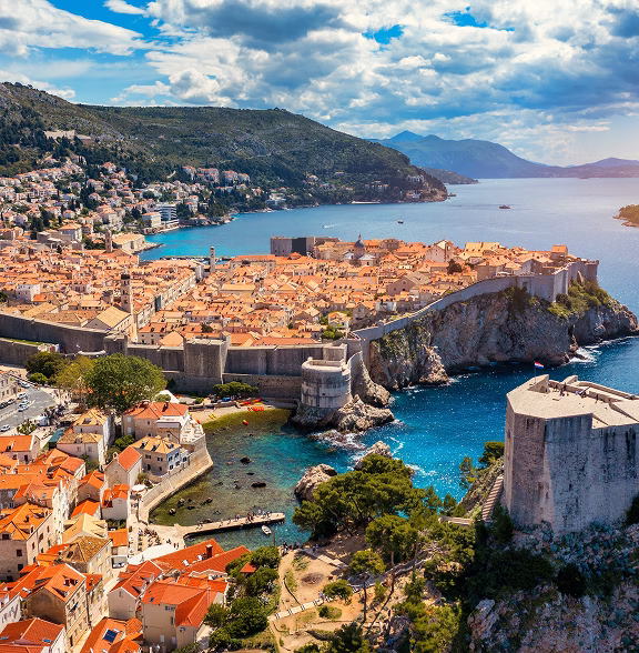 Aerial view of a fortified coastal city with orange rooftops, surrounded by clear blue water and green hills under a partly cloudy sky.